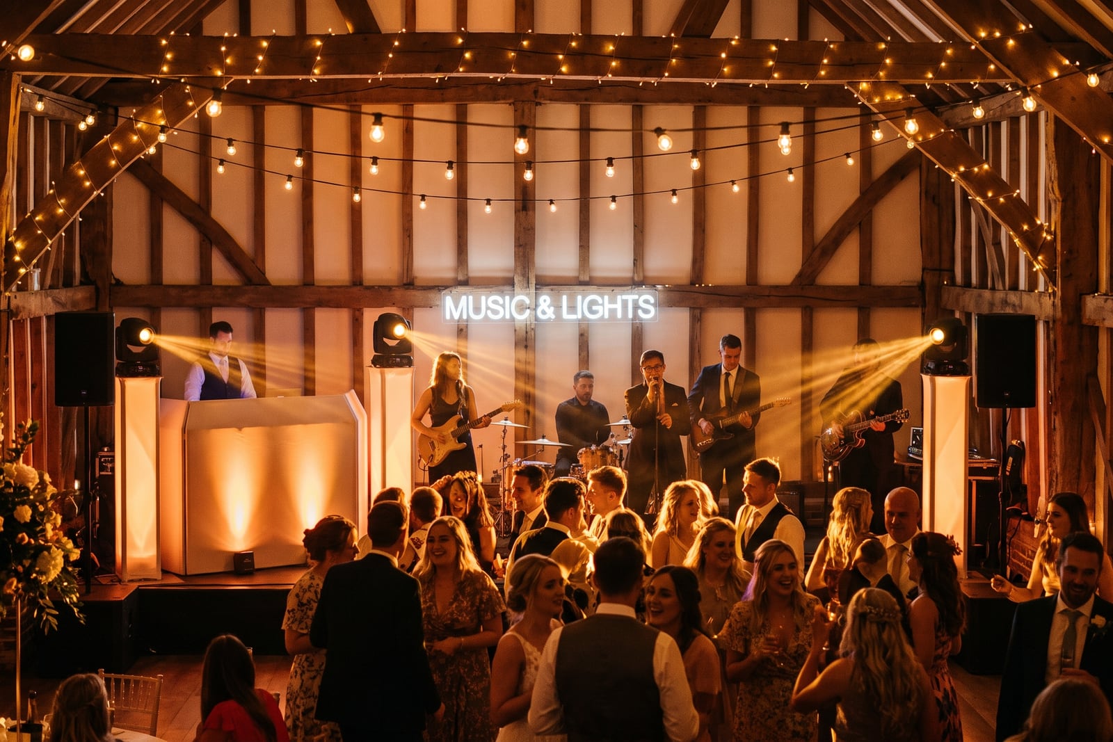 Live band and DJ on a barn stage under warm string lights and spotlights at a wedding reception